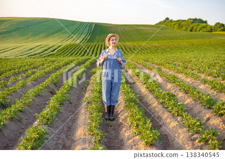 A young woman wearing overalls and a straw hat stands in a field of young crops with rolling green hills in the background A young woman wearing overalls and a straw hat stands in a field of young crops with rolling green hills in the background 138340545