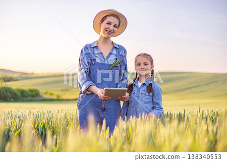 A woman in overalls and a straw hat holds a tablet while standing with her daughter in a sunlit wheat field, representing modern agriculture and family A woman in overalls and a straw hat holds a tablet while standing with her daughter in a sunlit wheat field, representing modern agriculture and family 138340553