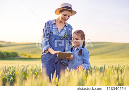 A woman in overalls and a straw hat and a young girl in denim look at a tablet computer in a sunlit wheat field 138340554