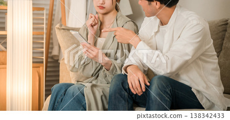 A young Asian couple is looking at their smartphones on the living room sofa, troubled and worried. A young Asian couple is looking at their smartphones on the living room sofa, troubled and worried. 138342433