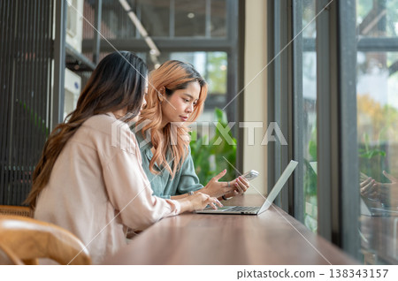 Smart asian woman holding phone looking at a friend's laptop while meetup for work or study in cafe. 138343157