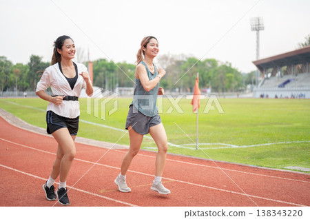 Full body of athletic asian woman and friend in sportswear running together on racetrack in stadium. 138343220