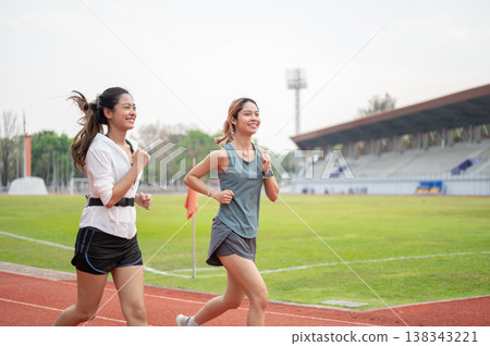 Happy athletic asian woman and friend in sportswear running jogging together on racetrack in stadium Happy athletic asian woman and friend in sportswear running jogging together on racetrack in stadium 138343221