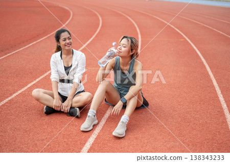 Asian woman sitting on racetrack aside a friend drinking water while resting after running exercise Asian woman sitting on racetrack aside a friend drinking water while resting after running exercise 138343233