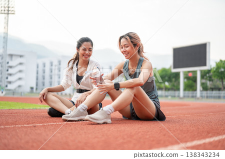 Asian woman holding water bottle talking to friend while sitting on racetrack resting after running. Asian woman holding water bottle talking to friend while sitting on racetrack resting after running. 138343234