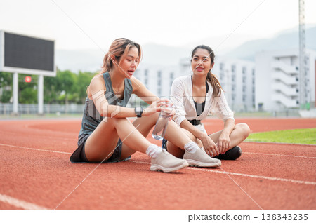 Asian woman holding water bottle talking to a friend as sitting on racetrack resting after running. Asian woman holding water bottle talking to a friend as sitting on racetrack resting after running. 138343235