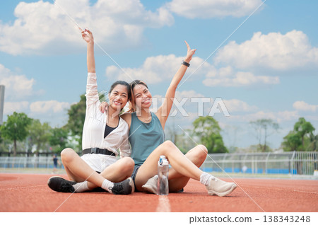 Happy asian woman hugging a friend's shoulder doing v peace hand sign while sitting on running track Happy asian woman hugging a friend's shoulder doing v peace hand sign while sitting on running track 138343248