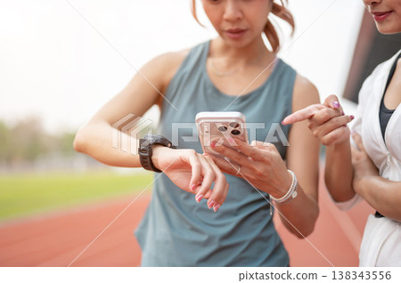 Close up of woman friends checking time on phone and watch while exercise on track in sports stadium Close up of woman friends checking time on phone and watch while exercise on track in sports stadium 138343556