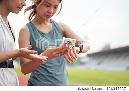 Close up of asian woman friends checking time on phone and watch while exercising in sports stadium. Close up of asian woman friends checking time on phone and watch while exercising in sports stadium. 138343557