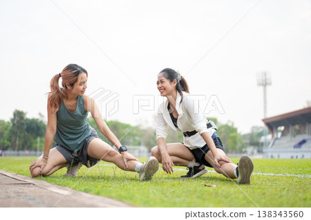 Asian woman and friend stretching leg muscles before exercise on green grass field in sports stadium 138343560