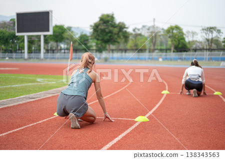 Back view of athlete woman and friend sitting crouching to start running on track in sports stadium. 138343563