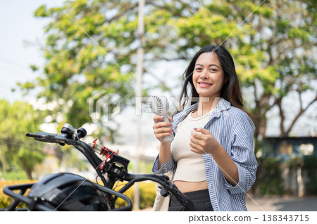 Pretty asian woman holding portable fan enjoy cooling wind while standing with black bicycle in park Pretty asian woman holding portable fan enjoy cooling wind while standing with black bicycle in park 138343715