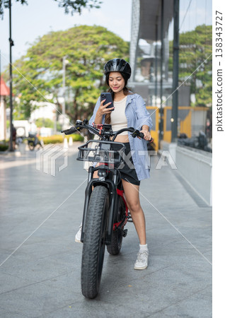 Full body asian woman wearing helmet looking at phone while riding e bike electric bicycle in park. Full body asian woman wearing helmet looking at phone while riding e bike electric bicycle in park. 138343727