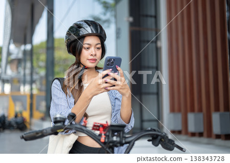 Pretty asian woman wearing helmet looking at phone while stopped riding a bicycle aside a building. Pretty asian woman wearing helmet looking at phone while stopped riding a bicycle aside a building. 138343728