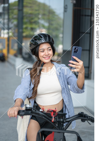 Pretty asian woman wearing helmet looking at phone taking a photo while riding on a bicycle in park. Pretty asian woman wearing helmet looking at phone taking a photo while riding on a bicycle in park. 138343730