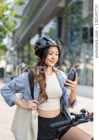 Pretty asian woman wearing helmet carrying eco bag looking at phone while riding a bicycle in park. 138343731