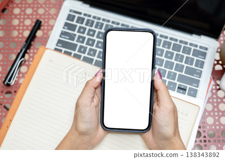Top view of a woman's hands holding white screen smartphone over book and laptop at table in cafe. Top view of a woman's hands holding white screen smartphone over book and laptop at table in cafe. 138343892
