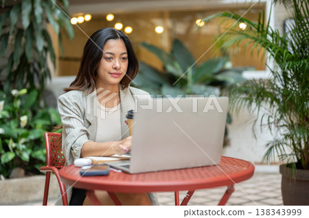 Pretty asian businesswoman holding coffee working on a laptop while sitting at table in garden cafe. Pretty asian businesswoman holding coffee working on a laptop while sitting at table in garden cafe. 138343999