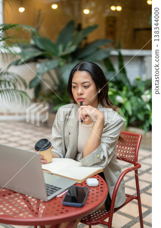 Pretty asian businesswoman holding pen thinking as looking at laptop sitting at table in garden cafe Pretty asian businesswoman holding pen thinking as looking at laptop sitting at table in garden cafe 138344000