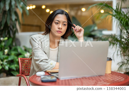 Pretty asian businesswoman holding pen touching head looking at laptop sits at table in garden cafe. Pretty asian businesswoman holding pen touching head looking at laptop sits at table in garden cafe. 138344001