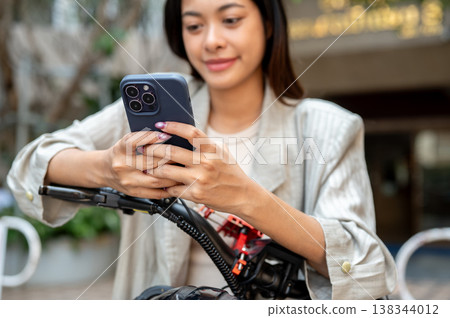 Close up of woman holding smartphone with both hands riding standing over bicycle in front of condo. 138344012