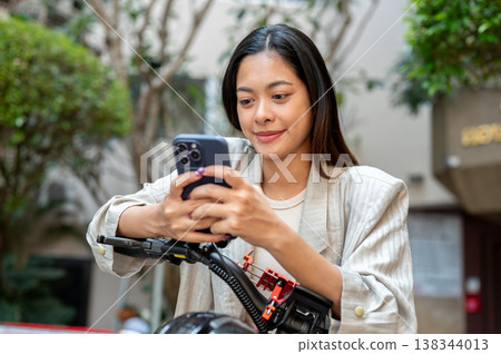 Happy asian woman looking at phone holding both hands riding standing over bicycle in front of condo Happy asian woman looking at phone holding both hands riding standing over bicycle in front of condo 138344013