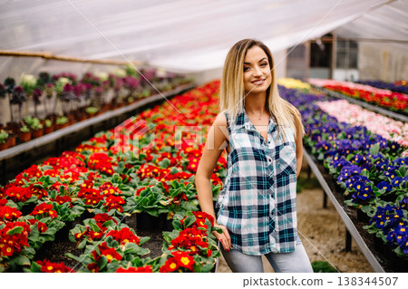 Women gardener in botanic garden,planting flowers and decorate.Smiling happy girl work in the garden.Young woman with scarf and cap kneeling between blooming plants Women gardener in botanic garden,planting flowers and decorate.Smiling happy girl work in the garden.Young woman with scarf and cap kneeling between blooming plants 138344507
