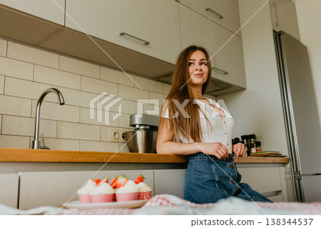 Portrait of friendly smiling female professional confectioner topping a cupcake with cream using a pastry bag. Looking at the camera. Indoors image. Pastry chef woman making creamy cakes Portrait of friendly smiling female professional confectioner topping a cupcake with cream using a pastry bag. Looking at the camera. Indoors image. Pastry chef woman making creamy cakes 138344537