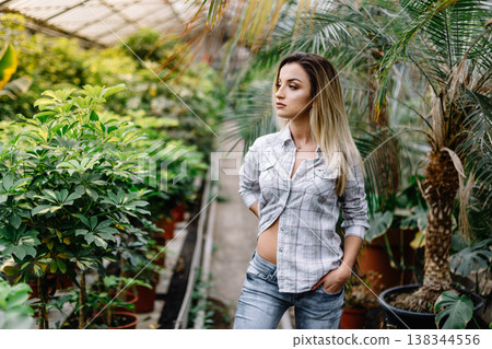 Smiling happy Caucasian female florist worker holding in crate flowers while standing in greenhouse. Smiling girl in the garden center. Smiling employee in garden center. 138344556