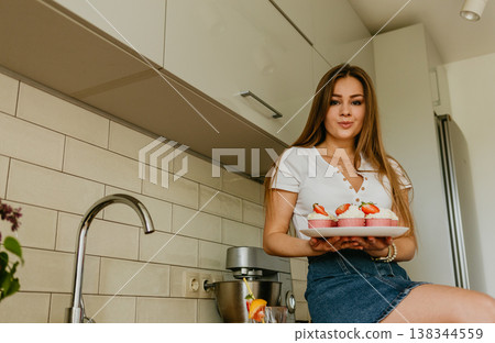 Portrait of friendly smiling female professional confectioner topping a cupcake with cream using a pastry bag. Looking at the camera. Indoors image. Pastry chef woman making creamy cakes. 138344559