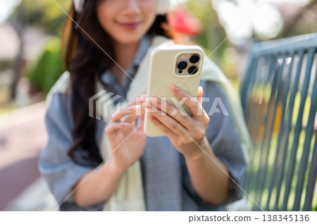 Close up of a woman wearings headphones looking at smartphone while sitting on bench in garden park. 138345136