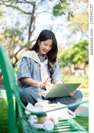 Pretty asian woman waving a hand at laptop while sitting folding legs on bench in the garden or park 138345137