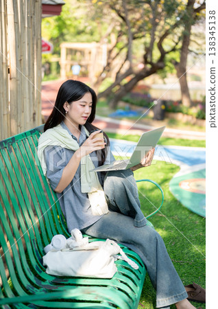 Pretty asian woman holding coffee watching on laptop while sitting knee up on bench in garden park. 138345138