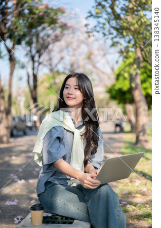 Pretty asian woman looking away from laptop while sitting cross legged on concrete bench at sidewalk Pretty asian woman looking away from laptop while sitting cross legged on concrete bench at sidewalk 138345140