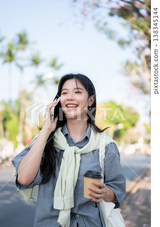 Smiling asian woman holding coffee cup talking on phone call while standing or walking by the road. 138345144