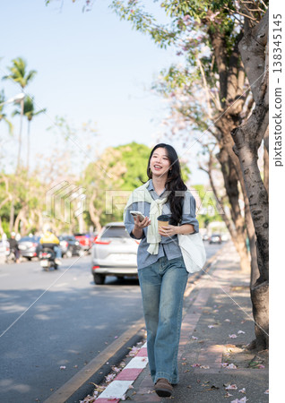 Full body of a pretty asian woman holding coffee cup and phone while walking on sidewalk by the road Full body of a pretty asian woman holding coffee cup and phone while walking on sidewalk by the road 138345145