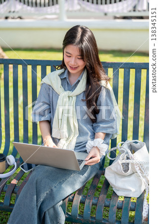 Pretty asian woman looking at a laptop holding on crossed legs while sitting on bench in garden park Pretty asian woman looking at a laptop holding on crossed legs while sitting on bench in garden park 138345151