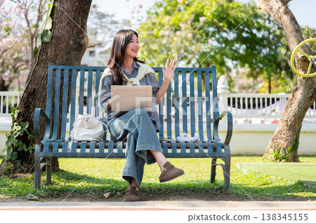Pretty asian woman waving a hand over laptop while sitting cross legged on bench in the garden park. Pretty asian woman waving a hand over laptop while sitting cross legged on bench in the garden park. 138345155