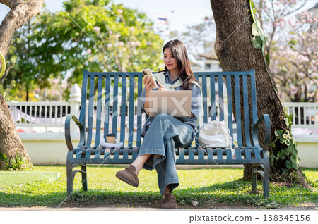 Pretty asian woman looking at phone over laptop while sitting cross legged on bench in garden park. Pretty asian woman looking at phone over laptop while sitting cross legged on bench in garden park. 138345156