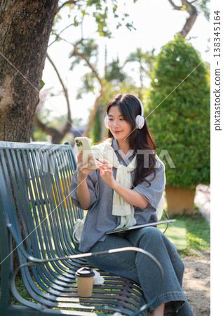 Pretty asian woman listening to music as looking at smartphone while sitting on bench in garden park 138345164