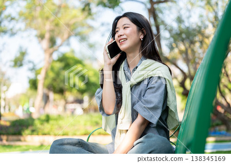 Pretty asian woman smiling as talking on phone while sitting folding legs on bench in garden or park 138345169