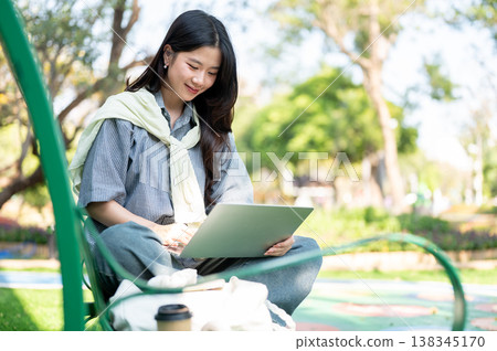 Pretty asian woman looking at laptop over folding legs while sitting on bench in the garden or park. 138345170