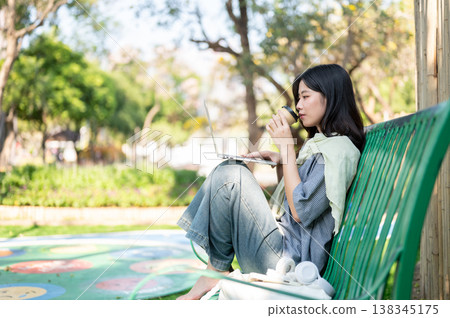 Pretty asian woman drinking coffee looking at laptop on knees while sitting on bench in garden park. Pretty asian woman drinking coffee looking at laptop on knees while sitting on bench in garden park. 138345175