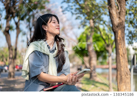 Happy asian woman holding phone and book looking away while sitting on bench at sidewalk by the road 138345186