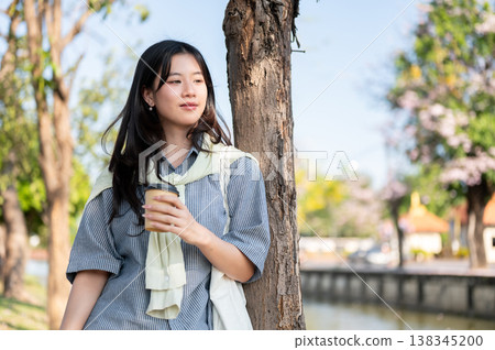 Pretty asian woman holding coffee enjoy the scenery while standing leaning on a tree by city's canal 138345200