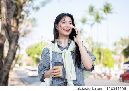 Happy asian woman holding coffee cup and talking on phone call while standing or walking by the road Happy asian woman holding coffee cup and talking on phone call while standing or walking by the road 138345206