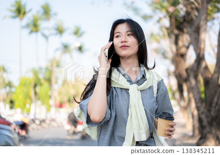 Pretty asian woman holding coffee taking or making a phone call while standing or walking by a road. Pretty asian woman holding coffee taking or making a phone call while standing or walking by a road. 138345211