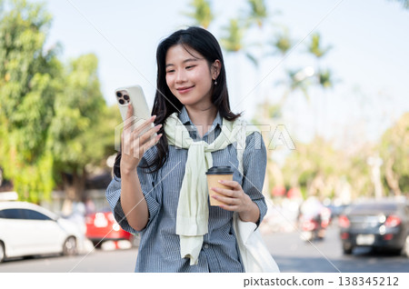 Smiling asian woman holding coffee cup looking at smartphone while standing or walking by the road. 138345212