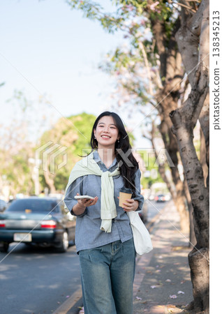 Asian woman smiling happily as holding coffee cup and phone while walking on sidewalk by the road. Asian woman smiling happily as holding coffee cup and phone while walking on sidewalk by the road. 138345213
