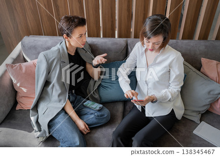 Female coworkers in casual business clothing talking candidly together on sofa in modern open office. Coworkers, business, candid, sofa, teamwork, office. Female coworkers in casual business clothing talking candidly together on sofa in modern open office. Coworkers, business, candid, sofa, teamwork, office. 138345467